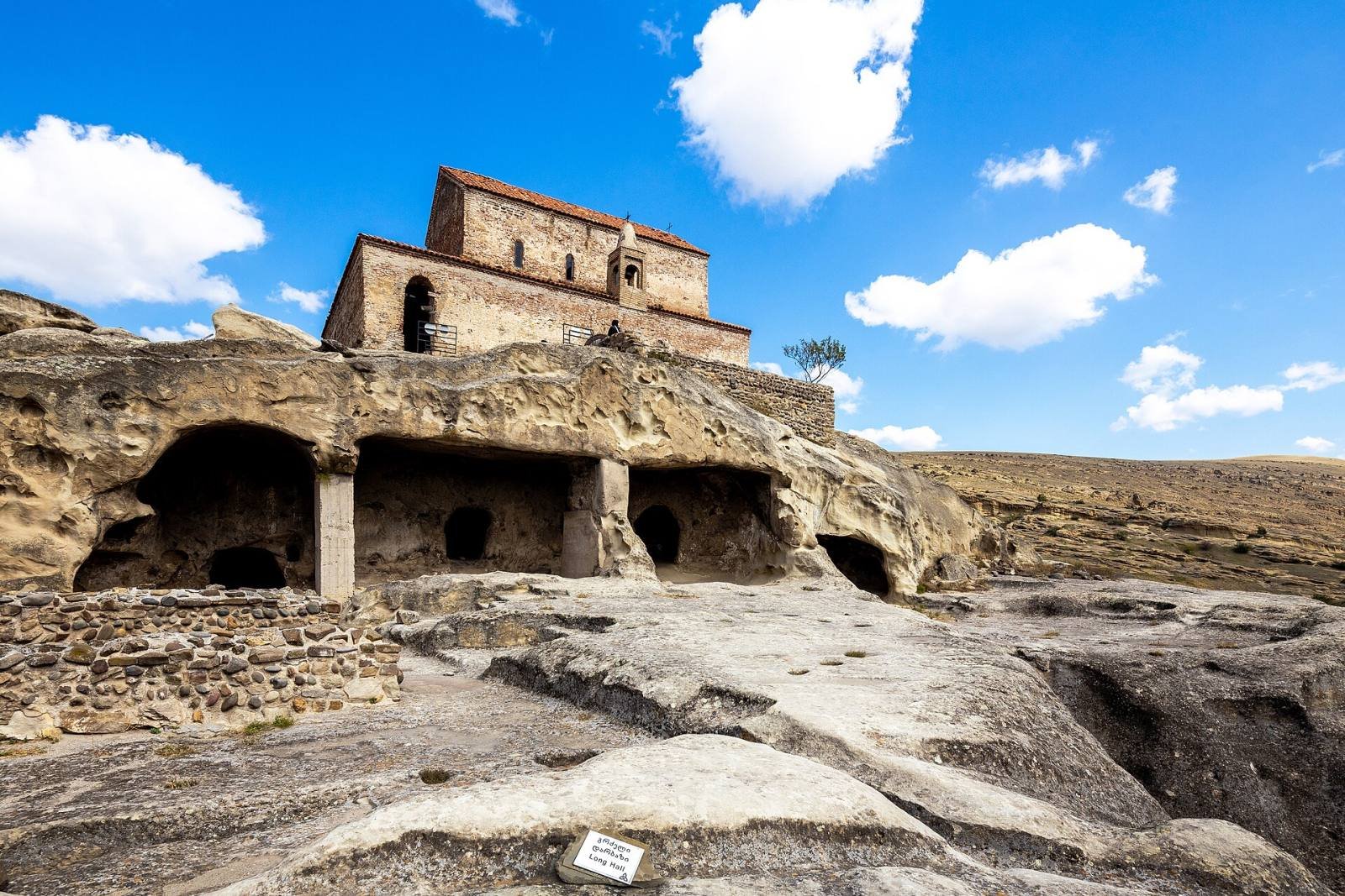 A medieval church of brick and stone standing on top of the cave dwellings of Uplistsikhe under a blue sky