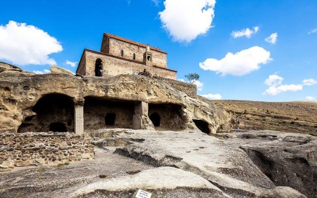 A medieval church of brick and stone standing on top of the cave dwellings of Uplistsikhe under a blue sky