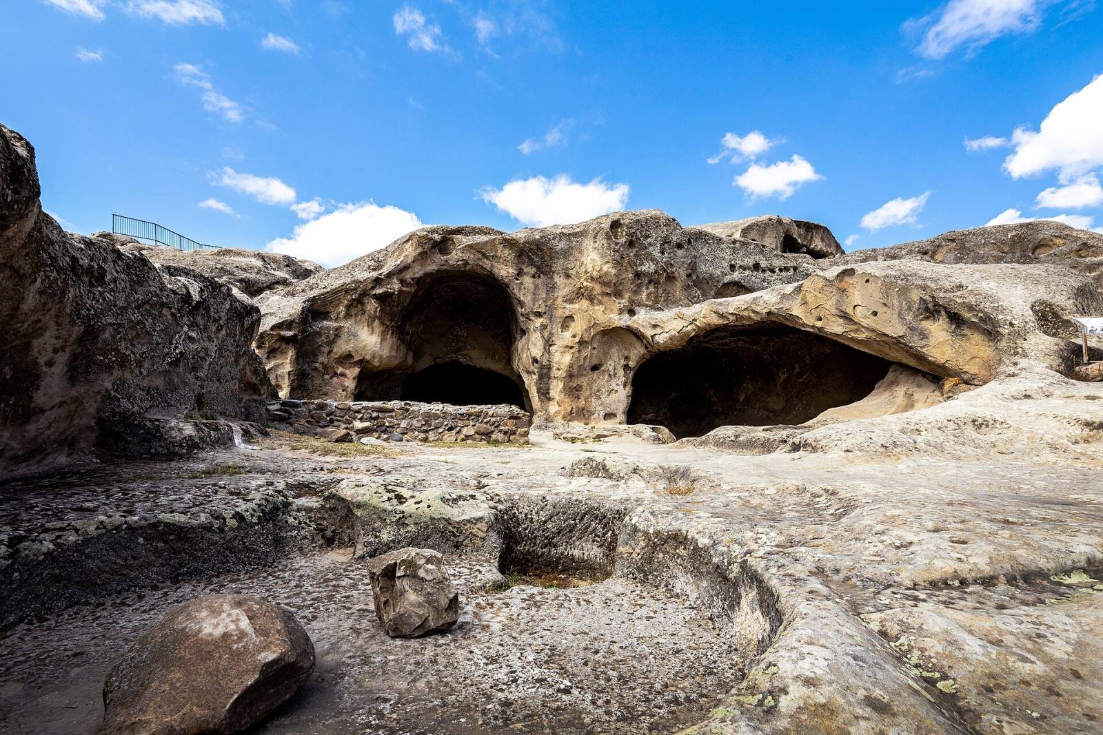 Large cave halls cut into the pale sandstone at Uplistsikhe under a blue sky with white clouds