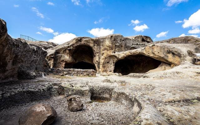Large cave halls cut into the pale sandstone at Uplistsikhe under a blue sky with white clouds